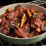 Braised lamb shanks in a pan with rosemary sprigs, pomegranate seeds, and visible sauce, cooking in the oven.