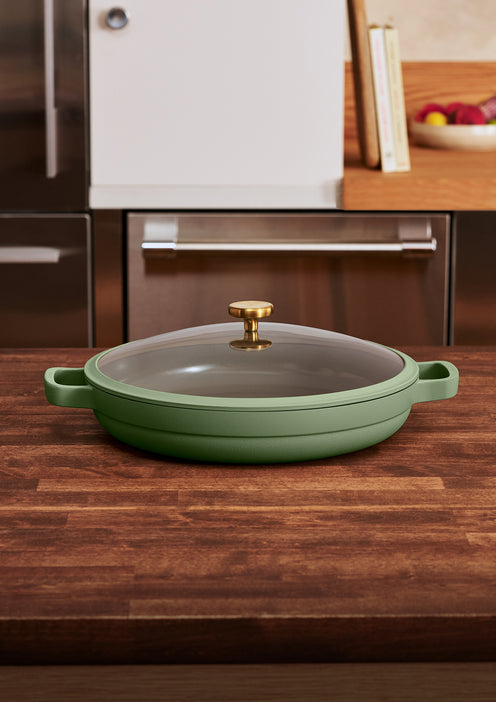 A green pan with a glass lid and gold handle sits on a wooden kitchen counter. Kitchen cabinets and a fruit bowl are visible in the background.