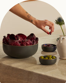 A hand with a tattoo reaches for a black bowl of Kalamata olives on a table. The table also holds a black bowl of green olives, a large black bowl of radicchio, a ceramic pitcher, and a spiky blue flower in a vase.