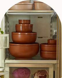 A refrigerator shelf filled with neatly stacked, matching brown mixing bowls of various sizes, with purple cabbage and other vegetables visible in the bottom drawer.