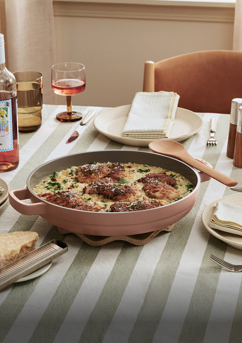 A skillet of cooked chicken and rice sits on a striped tablecloth, surrounded by plates, napkins, cutlery, a glass of rosé wine, and a bottle.