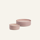 Two stacks of pink ceramic plates with brown rims are arranged on a plain light background: one stack is the Small Plates Set of four, and the other stack has four larger plates.