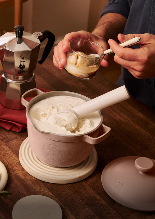 Person adding cream to a pot of coffee on a wooden table with a Stovetop espresso maker.