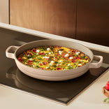 A baked frittata with vegetables and cheese sits in a round beige pan with handles on a modern stovetop.