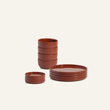 The Plates & Bowls Set features reddish-brown ceramic pieces—four small bowls, four plates, and four small saucers—neatly stacked against a plain off-white background.
