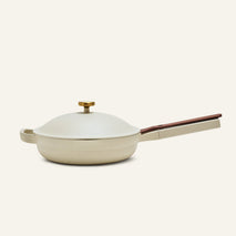 The Always Pan 2.0, a beige nonstick frying pan from a bestselling cookware set, comes with a matching domed lid, wooden handle, and brass knob, shown against a plain light background.