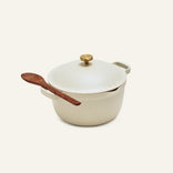 The Perfect Pot, a cream ceramic family-sized pot with a lid and gold knob, features a wooden spoon resting on its handle, shown against a plain light background.